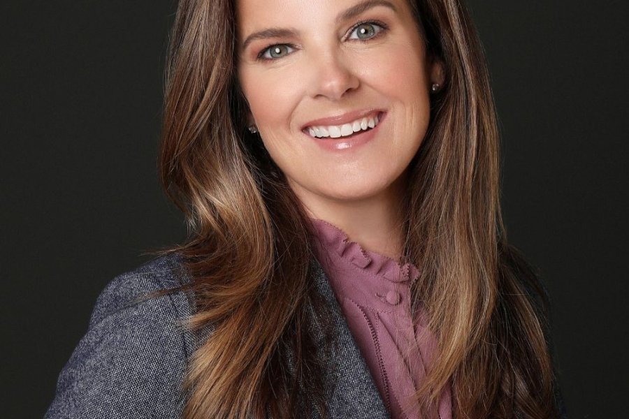Woman with long brown hair and a warm smile, wearing a gray blazer over a purple blouse, posed against a dark background.